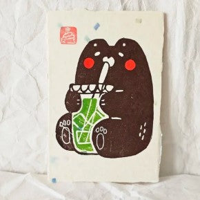 Bear Matcha Small Lino Print by Misu Juju from Blue Dog Posters features a cute black bear with red cheeks sipping an iced green drink through a straw, sitting on a textured white surface in Misu Juju's signature lino print style.