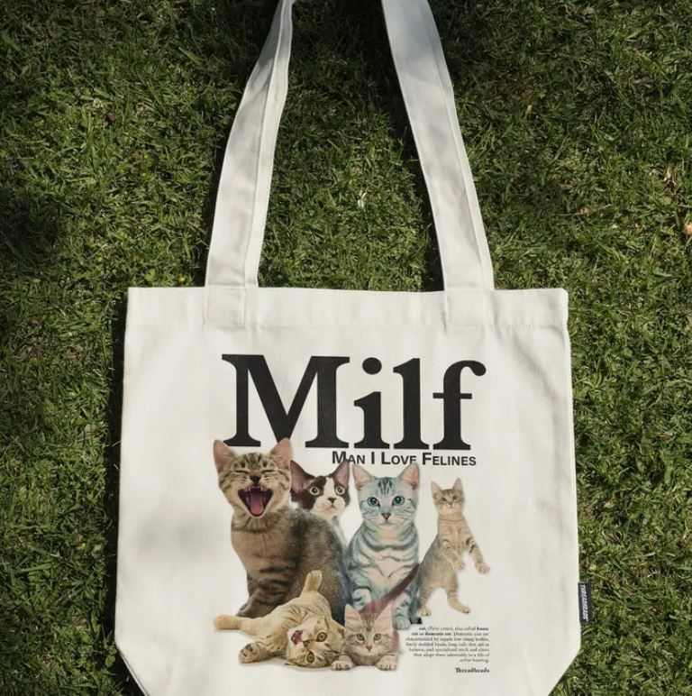 A natural cotton canvas tote bag by Blue Dog Posters rests on grass, showing five cats and bold "Milf" text with the playful caption "Man I Love Felines" beneath. Product: Man I Love Felines Tote Bag Natural.