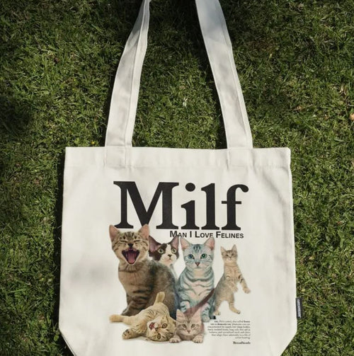 A natural cotton canvas tote bag by Blue Dog Posters rests on grass, showing five cats and bold "Milf" text with the playful caption "Man I Love Felines" beneath. Product: Man I Love Felines Tote Bag Natural.