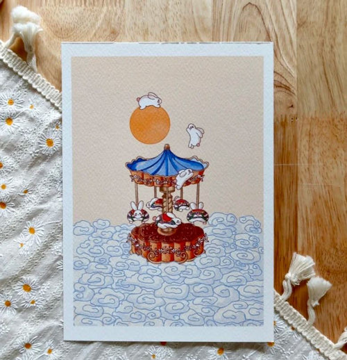 Mooncake Carousel" by Plain Rice Studios for Blue Dog Posters shows rabbits riding a blue-orange carousel under an orange sun, with swirling clouds. The digital art postcard is displayed on wood with a white embroidered cloth.