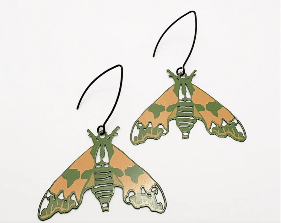 The Moth Lime Hawk Earrings by Denz, from Blue Dog Posters, feature green and tan camouflage moth shapes with black hooks. Crafted from recycled stainless steel, they're shown against a white background.