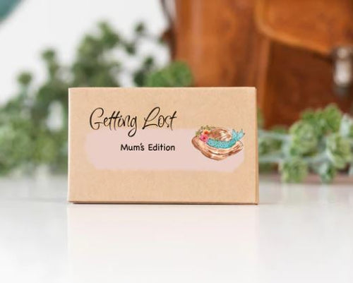 A small brown box labeled "Getting Lost Cards - Mums Edition" from Blue Dog Posters, featuring a colorful illustration of a nest and eggs. Perfect for Mother's Day, this travel adventure game is placed on a white surface with green plants and a brown leather bag in the blurred background.