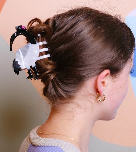 A woman with brown hair in a bun, secured with Blue Dog Posters’ Ibis Bin Chicken Hair Claw made from plant-based acetate, wears gold hoops and a lavender top. A pastel background lends softness to the scene.