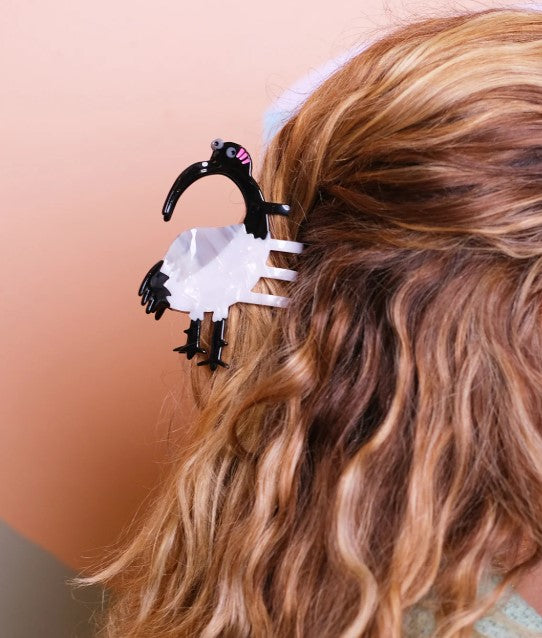 A person with wavy, light brown hair wears the Blue Dog Posters Ibis Bin Chicken Hair Claw, a black and white bird-shaped clip made from plant-based acetate, attached to the side of their head against a peach-colored background.