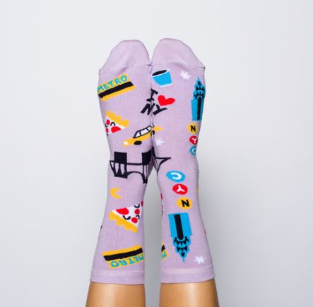 Two feet are shown wearing Blue Dog Posters’ New York Womens Socks—purple cotton crew socks featuring NYC-themed designs like pizza, a taxi, skyscrapers, coffee, metro signs, and "I ♥ NY"—against a plain white background.