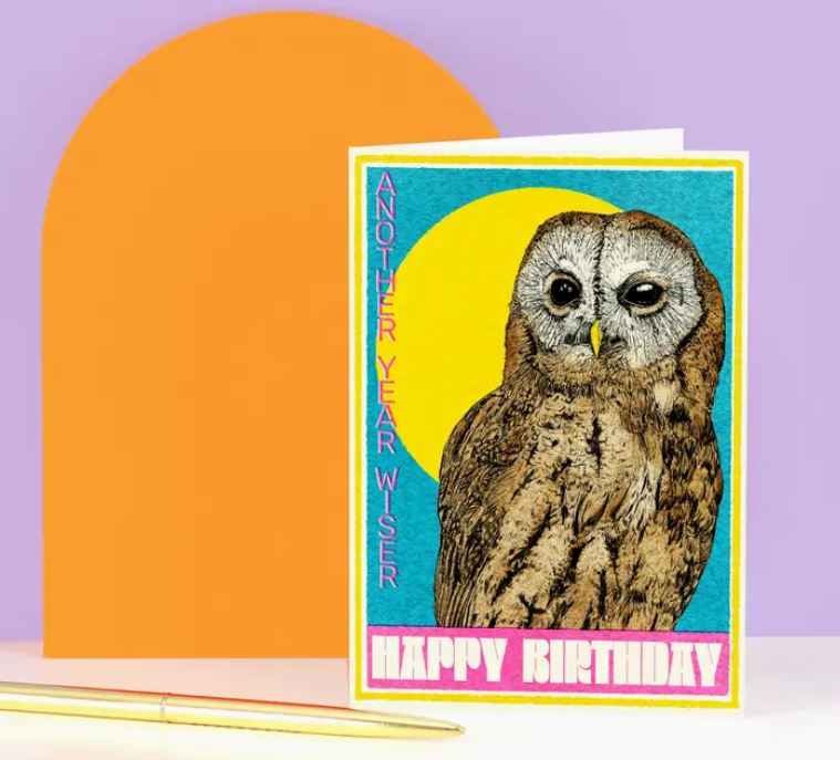 On a table sits the "Another Year Wiser Owl Birthday Card" by Blue Dog Posters, featuring an owl illustration with the words "ANOTHER YEAR WISER" and "HAPPY BIRTHDAY," set against a blue sky with a sun-like yellow circle, alongside a pen and orange semi-circle.