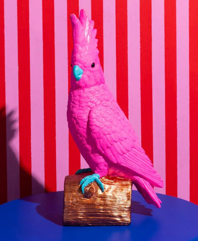 Pablo The Parrot Hot Pink Resin Animal by Blue Dog Posters features bright pink feathers, a turquoise beak and feet, and sits on a gold perch—ideal for colorful décor. Set against red and pink stripes with a solid blue base, it’s truly eye-catching.
