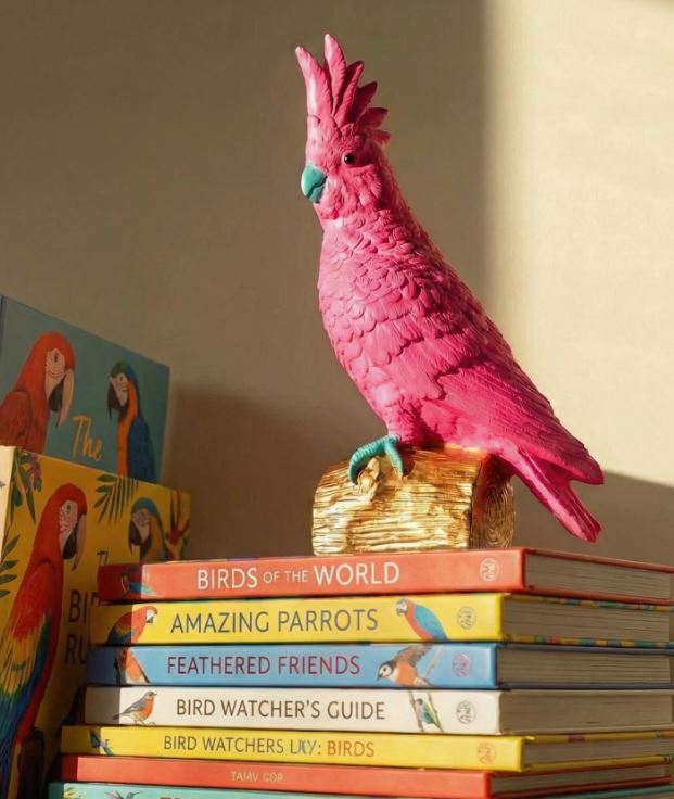 The Pablo The Parrot Hot Pink Resin Animal by Blue Dog Posters perches on a gold base atop vibrant bird books, adding a cheerful pop of colour to your decor as sunlight casts gentle shadows.