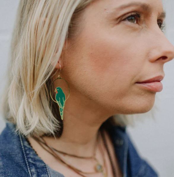 A woman with short blonde hair wears Parrot in Green Denz Earrings by Blue Dog Posters, layered necklaces, and a denim jacket—her look exudes subtle Australiana flair.