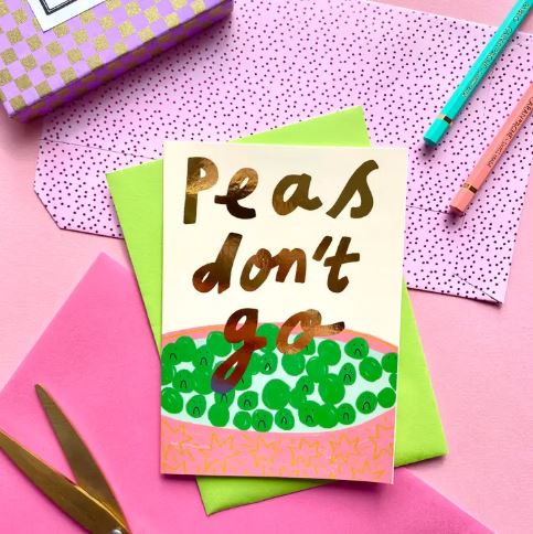 The "Peas Don't Go Greeting Card" by Blue Dog Posters is an A6 FSC certified blank card with "Peas don't go" in gold above green peas, shown on pink with matching envelopes, scissors, pens, and a patterned box.
