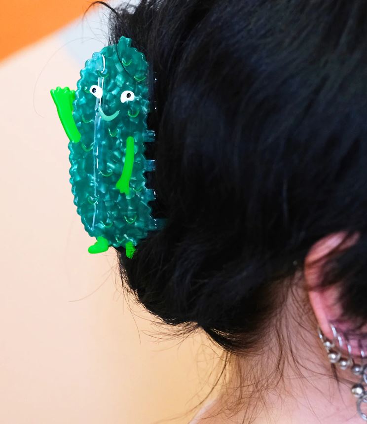 A close-up of dark hair clipped with the Pickle Hair Claw by Blue Dog Posters, a green plant-based acetate hair accessory shaped like a smiling cactus, with raised arms and part of the ear and earrings visible against a blurred background.