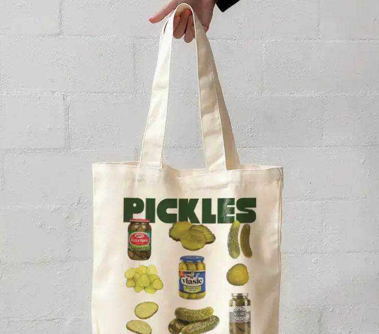 A hand holds The Pickles Tote Bag Natural by Blue Dog Posters, featuring images of pickles, jars, and slices with "PICKLES" in bold green letters at the top.