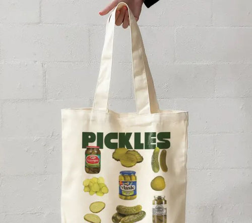 A hand holds The Pickles Tote Bag Natural by Blue Dog Posters, featuring images of pickles, jars, and slices with "PICKLES" in bold green letters at the top.