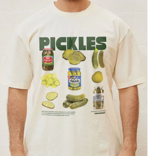 Someone wearing The Pickles T-Shirt Black LGE by Blue Dog Posters, featuring bold green "PICKLES" lettering and playful pickle-themed graphics with slices, jars, whole pickles, and brands like Vlasic and Bubbies printed below.