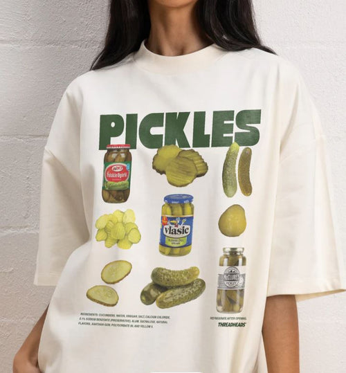 A person sports The Pickles T-Shirt White MED by Blue Dog Posters, featuring bold green "PICKLES" text and graphic pickles for a fun, retro vibe.
