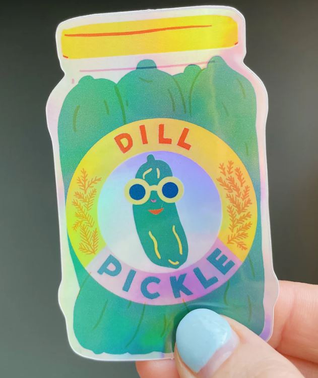 The Pickle Sticker by Blue Dog Posters features a holographic "Dill Pickle" jar with a smiling cartoon pickle in sunglasses at the center, designed by Bristol illustrator Naomi Wilkinson and shown held by a hand with light blue nails.