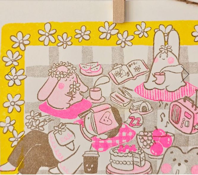 Picnic Riso Print by Misu Juju from Blue Dog Posters features cute cartoon bunnies on a checkered picnic blanket, surrounded by snacks, books, flowers, and cozy items. The pink, yellow, and brown daisy border completes this charming A4 art print.