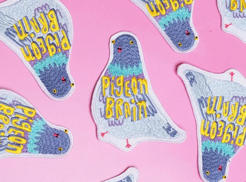 The Pigeon Brain Patch by Blue Dog Posters features "Pigeon Brain" embroidered in yellow on blue, gray, and green birds. These quirky iron-on patches are designed by Bea Bellingham and set against a playful pink background.