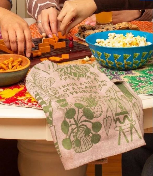 Three people play a board game at a round table with snacks. A Blue Dog Posters Plant Problem Dish Towel, decorated with green plant illustrations and the phrase “I have a bit of a plant problem,” hangs over the table’s edge.