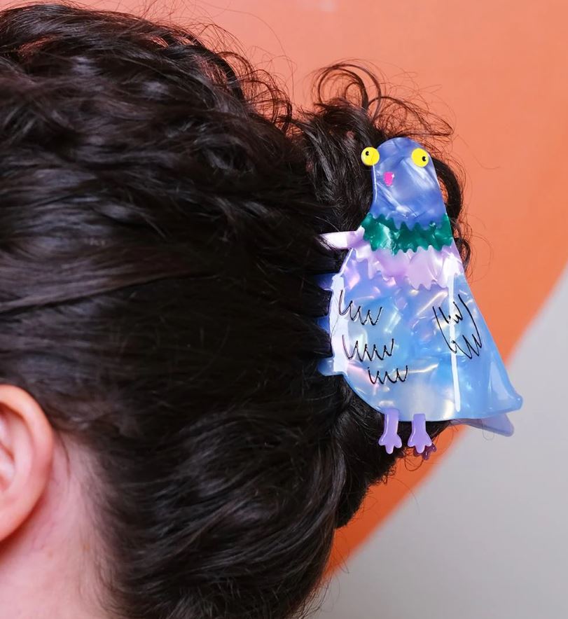 A close-up of dark curly hair in a bun, held by the Bird Brain Pigeon Hair Claw from Blue Dog Posters—an acetate bird clip with yellow eyes and a green collar—set against an orange and beige background.