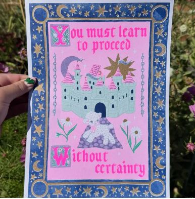 A hand holds the Blue Dog Posters "Proceed Without Certainty" A4 risograph by Amy Hastings, showing a castle, moon, stars, and flowers with an inspiring quote on recycled coffee cup paper, framed by a blue and gold celestial border.