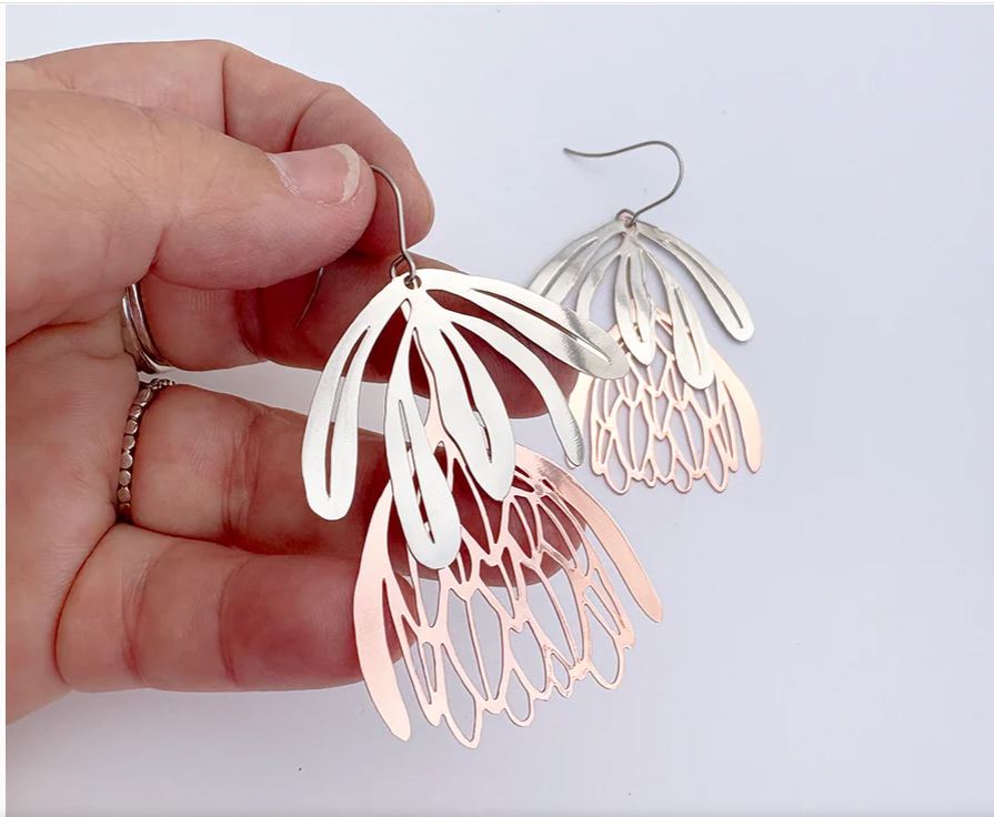 A hand holds Blue Dog Posters' Protea Flower Silver Rose Gold Denz Earrings—lightweight dangles with intricate, layered petal shapes in silver and rose gold; one earring rests on a white surface in the background.