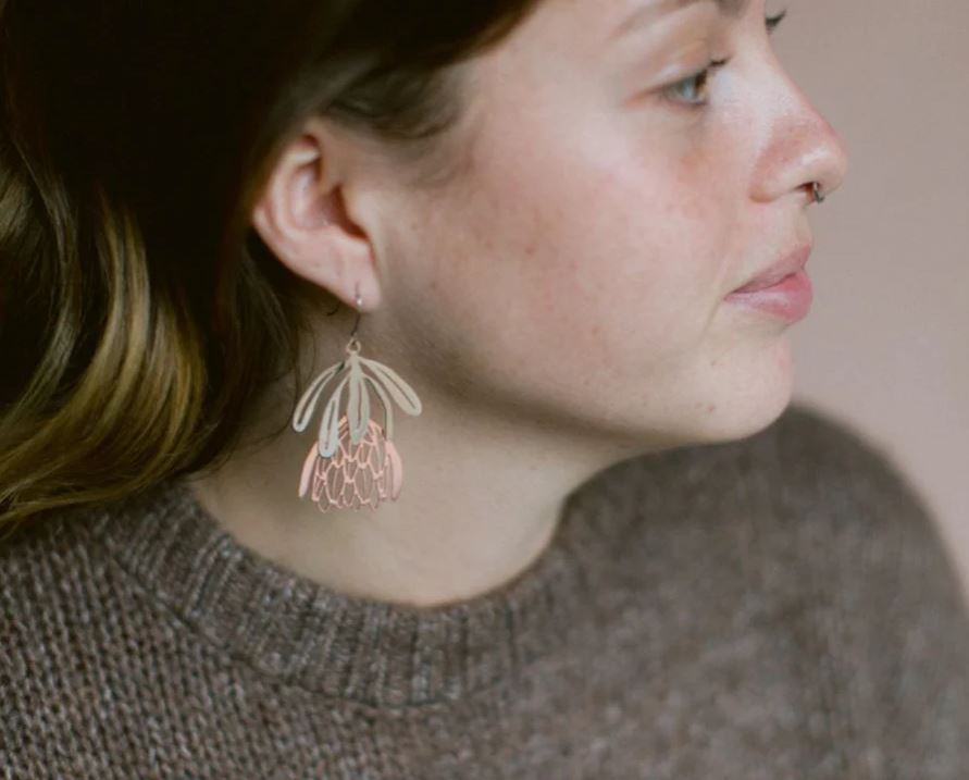 A light-skinned, brown-haired person wears a brown knit sweater and Blue Dog Posters' Protea Flower Silver Rose Gold Denz Earrings, shown in profile against a neutral background that highlights the delicate, nature-inspired Australian jewelry.