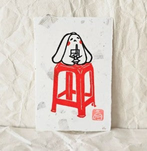 Bunny Red Chair (1) Small Lino Print by Misu Juju for Blue Dog Posters features a white, rabbit-like character with long ears sipping a drink on a tall red stool, inspired by lino print aesthetics and printed on textured white paper.