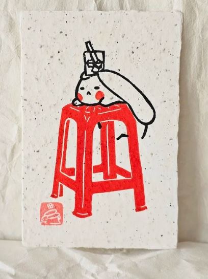 Bunny Red Chair (3) Small Lino Print by Misu-Juju from Blue Dog Posters features a minimalist, lino-inspired illustration of a faceless figure resting on a red chair with a drink atop its head, set against textured paper.