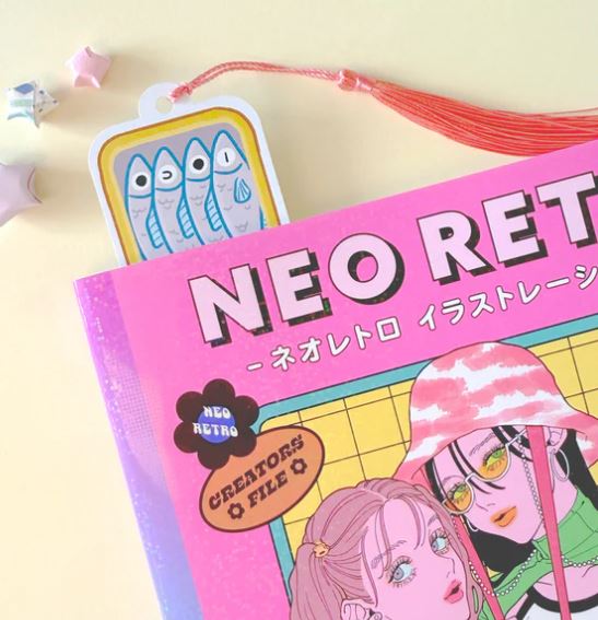 A pastel "NEO RETRO" book with two illustrated women in hats partly covers the Blue Dog Posters' Bookmark Sardine Stack, featuring blue fish. Origami stars and a red tassel add playful accents.