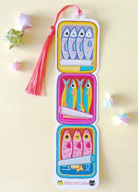 The Bookmark Sardine Stack by Blue Dog Posters is a quirky bookmark with three sections, each displaying illustrated sardines in blue, yellow, and pink cans. Features a pink tassel and playful origami stars.