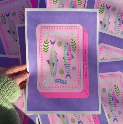A hand holds a colorful A4 Risograph illustration by Amy Hastings, featuring a pink sardine can with two fish and olives, branded "Sardines" in bold. The background is a purple stack of the same Blue Dog Posters design, creating a layered effect.