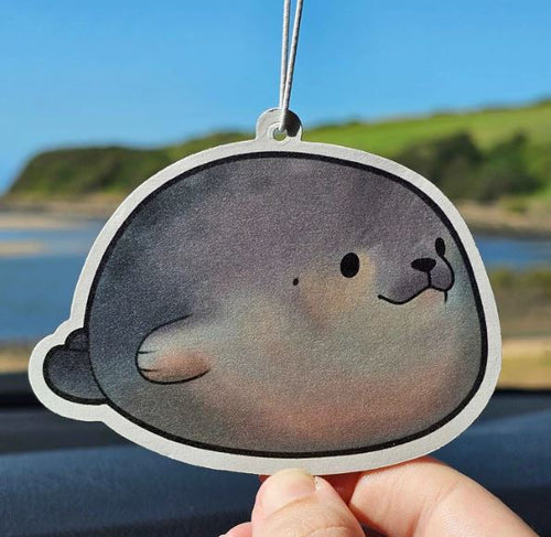 A hand holds up the Blue Dog Posters Round Seal Bubblegum Air Freshener - Pinstachiio, featuring a cute cartoon seal face, with water, hills, and blue sky blurred in the background.