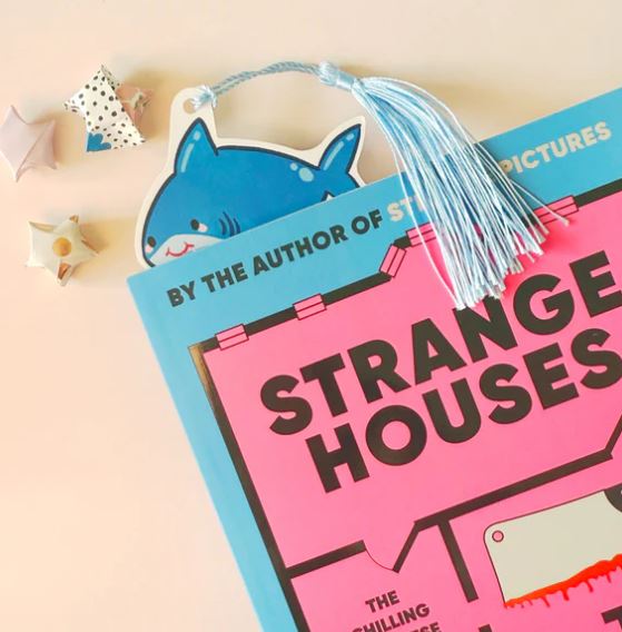 A book titled "Strange Houses" with a pink and black cover sits on a pale surface. The "Bookmark Shark Stack" by Blue Dog Posters, featuring a blue tassel, peeks out from the top beside three star-shaped stationery decorations.