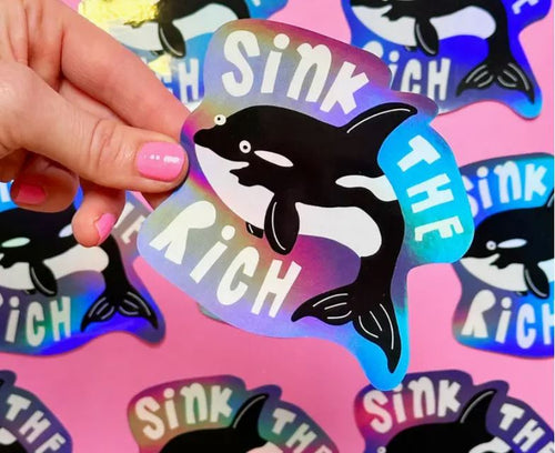 A hand with pink nail polish holds a Blue Dog Posters Sink the Rich Whale Holographic Sticker (100mm × 85mm) on a pink background covered in identical, water-resistant stickers.
