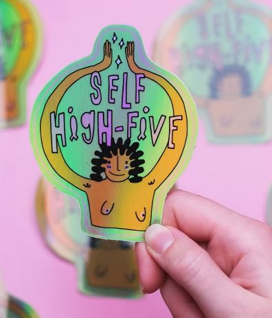 A hand holds a Blue Dog Posters Self High Five Holographic Sticker, showing a smiling figure raising their arms on a pink background with other vibrant stickers visible.