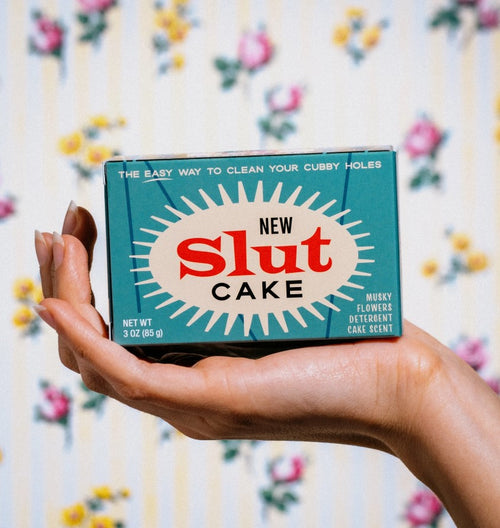 A hand holds a vintage-style teal box labeled "Slut Cake Triple Milled Boxed Bar Soap" in bold letters. The box also features text that reads "The easy way to clean your cubby holes" and "Musky flowers detergent cake scent." The background shows a floral wallpaper pattern, reminiscent of Blue Dog Posters' aesthetic.