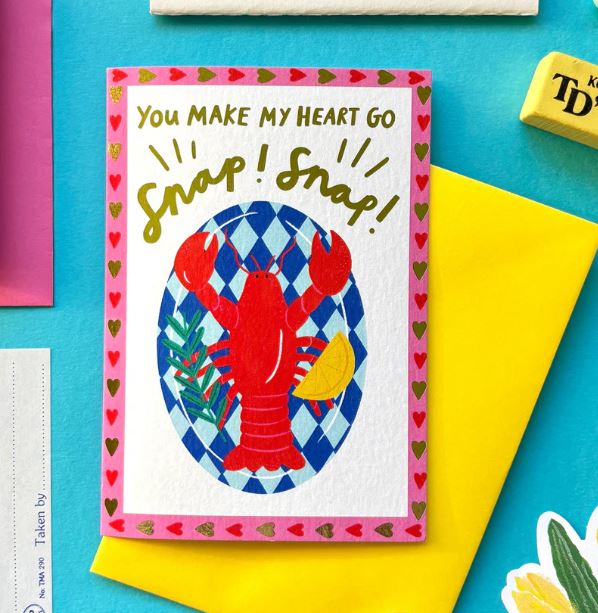 The You Make My Heart Go Snap Card by Blue Dog Posters features a red lobster, lemon wedge, green leaves on a blue patterned background, with "You make my heart go snap! Snap!" text and a pink border accented by green hearts.