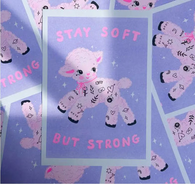 Illustration of a pastel purple lamb with cute tattoos, set against a blue background. Pink text above reads "Stay Soft," while the text below says "But Strong." Multiple pieces of the high quality A4 risograph print "Soft Strong" by Amy Hastings from Blue Dog Posters are arranged in a scattered pattern on 170gsm recycled coffee cup paper.