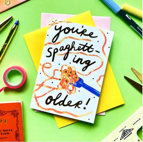 The "You're Spaghetting Older Greeting Card" by Blue Dog Posters features playful spaghetti art and a fun birthday pun, shown on a green desk surrounded by pens, markers, tape, and colored paper.