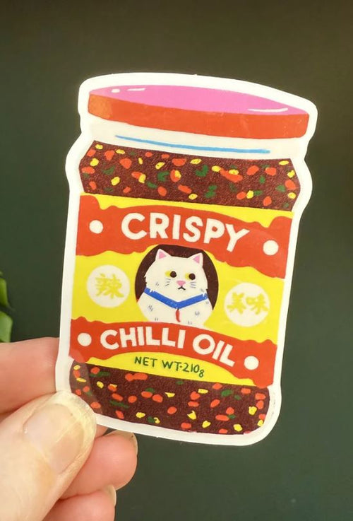 A hand holds the Crispy Chilli Oil Sticker by Blue Dog Posters, featuring Naomi Wilkinson’s design of a white cat and vibrant red, yellow, and orange chili flakes on a jar label.