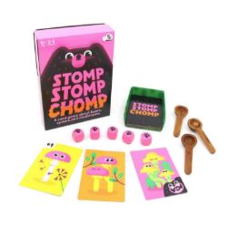 The Stomp Stomp Chomp card game set by Blue Dog Posters includes a pink box, colorful cards with cartoon mushrooms and bears, five pink dice with faces, a green tray, and five small wooden spoons, all displayed on a white background.
