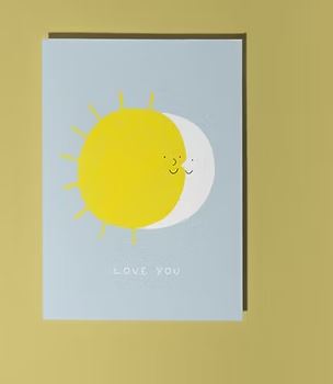 The Sun and Moon Love Card by Blue Dog Posters is a recyclable greeting card with an illustrated sun and moon hugging on a light blue background, "LOVE YOU" in white, perfect for Valentine’s Day. Card is shown resting on a yellow surface.