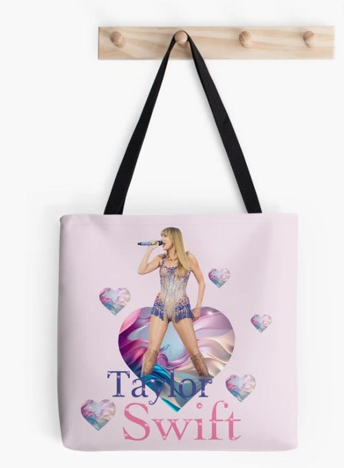 A Taylor Swift Hearts Tote Bag by Blue Dog Posters hangs on a wooden wall hook. The sublimation printed bag features an image of a woman with long blonde hair, singing into a microphone against a background of colorful, swirling patterns and heart shapes. The text reads "Taylor Swift" in blue and pink letters below the image from our Giftware Collection.