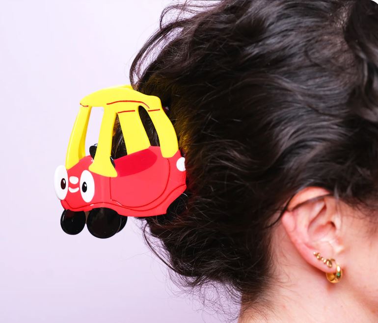 A person with dark hair wears the Tiny Tike Car Hair Claw by Blue Dog Posters, a playful cartoon car clip. Only the side of their head and gold earrings show against a light pink background, highlighting this nostalgic hair accessory.