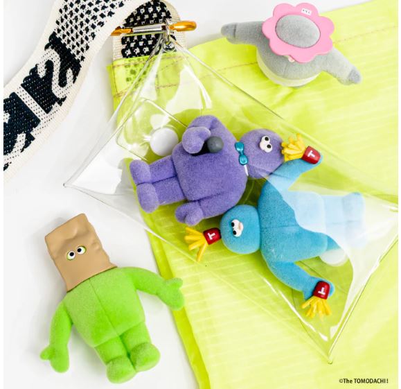 Four colorful plush toys from the Tomodachi Figure Vol 3 Blind Box by Blue Dog Posters, including a green square-headed figure and a blue bird, are displayed on yellow fabric with a clear pouch and white strap. A unique Japanese collectible set.