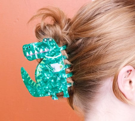Someone with light brown hair styles their bun using the T-Rex Hair Claw by Blue Dog Posters, a quirky green dinosaur-shaped accessory, against an orange background.