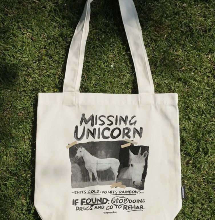 A cream cotton canvas tote bag by Blue Dog Posters lies on grass, featuring the "Missing Unicorn Tote Bag Natural" with a playful unicorn poster design and humorous rehab advice.