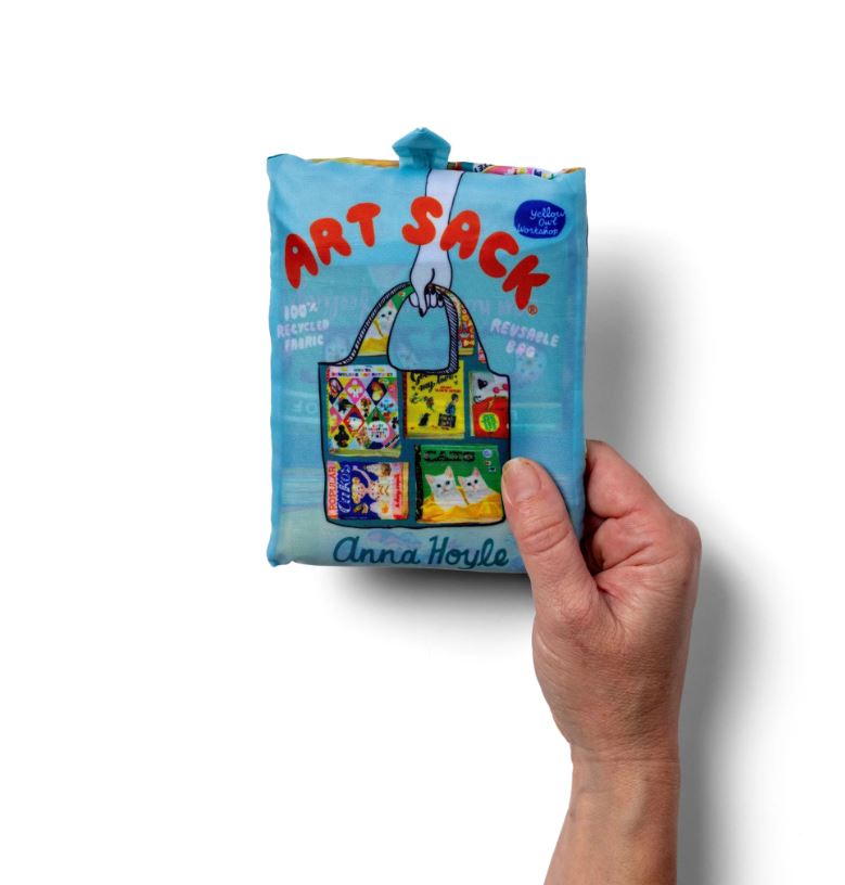 A hand holds a folded blue recycled rPET bag package labeled "Useless Books Art Sack" by Blue Dog Posters, featuring a colorful tote graphic and the text "Anna Hoyle" on the front against a white background.