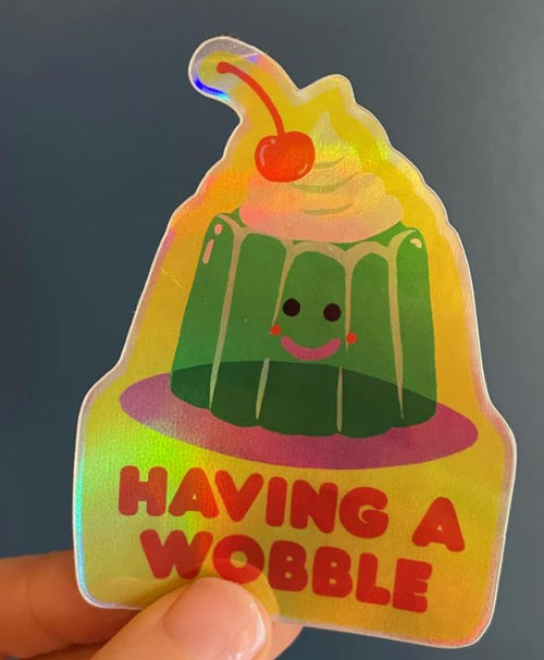 The Having a Wobble Sticker by Blue Dog Posters features Naomi Wilkinson’s holographic design of a smiling green jelly with a cherry, pink cheeks, and "HAVING A WOBBLE" in bold red at the bottom.